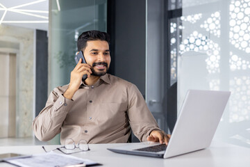 A professional young businessman chatting on the phone while working on his laptop in a modern office environment, conveying a sense of business communication and productivity.