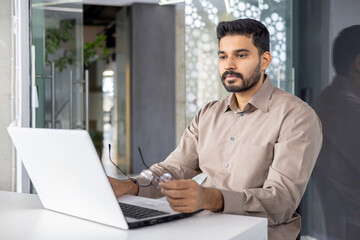 Obraz premium A focused professional man sits at a desk in a contemporary office, working intently on his laptop. His thoughtful expression and business casual attire reflect a typical workday environment.
