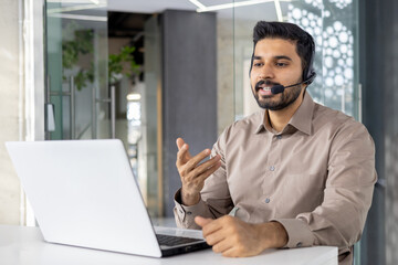 A young professional man engaging in customer service using a headset in a well-lit modern office. He gestures while speaking, emphasizing active communication.