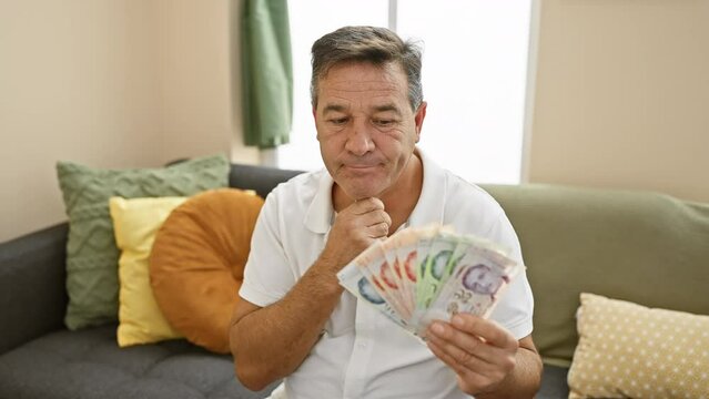 Shocked middle age man, covering mouth with hand holding singapore banknotes, afraid of a surprise accounting error in his homely living room