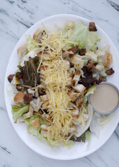 Caesar salad. Top view of fresh a salad with lettuce, parmesan cheese, bread croutons, sliced grilled chicken breast, bacon and Caesar dressing on the white marble table