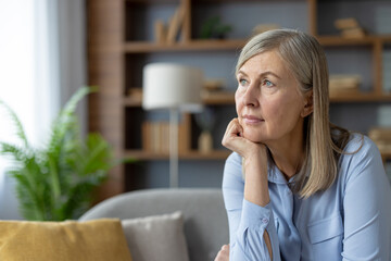 A serene mature woman in a blue shirt stares thoughtfully while sitting in a well-lit living room....