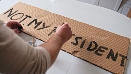 Caucasian Male Preparing Banner for Political Demonstration Raid before Presidential Election with Not My President Text Painted on Cardboard - obrazy, fototapety, plakaty