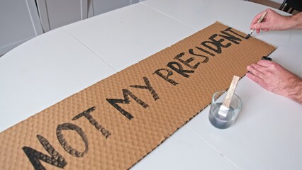 Caucasian Male Preparing Banner for Political Demonstration Raid before Presidential Election with Not My President Text Painted on Cardboard - obrazy, fototapety, plakaty