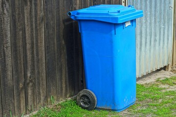 one plastic blue closed trash bin on wheels stands on the ground during the day near a wooden fence on the street
