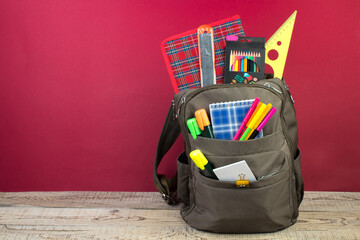 Backpack with different colorful stationery on table. Burgundy background. Back to school.