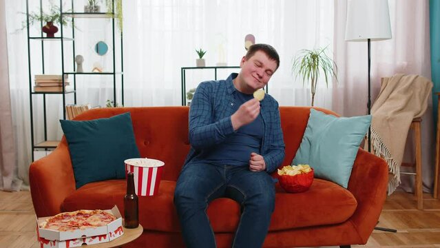 Happy Caucasian young man in casual clothes sitting on comfy sofa having chips, pizza, popcorn and beer watching movie sports enjoying weekend at home. Smiling guy eating junk food on couch in house.
