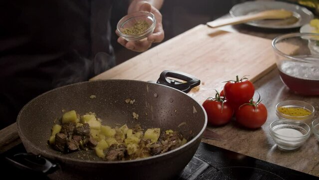 cook in the restaurant kitchen drizzles pan-fried meat with potatoes with a variety of spices. Tomatoes and other garnishes are beautifully arranged on the table, surrounding the dish. Close up. Slow