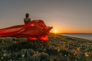 A woman in a red dress is standing in a field with the sun setting behind her. She is reaching up...