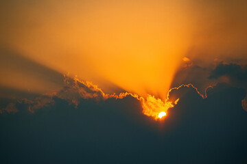 The sun is setting behind a cloud. The sky is orange and the clouds are dark. The sun is shining...