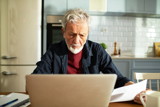 Senior man looking concerned while using laptop in kitchen with documents on table