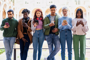Group of young adult students stands engrossed in their smartphones - connectivity and individual isolation - social network and technology concept - university courtyard background. - obrazy, fototapety, plakaty