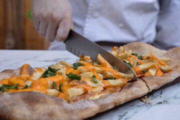 Closeup view of the chef holding a kitchen knife, slicing a pizza with fried potatoes, cheddar and mozzarella cheese, green onions, and bacon.