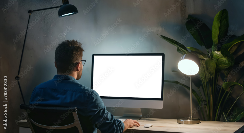 Poster a man is seated at a desk, focusing on the computer screen in front of him while working.