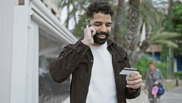 Smiling hispanic man talking on phone, holding card on city street.