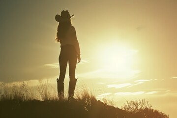 Naklejka premium Silhouetted woman in cowboy hat standing against sunset sky above grassy hill