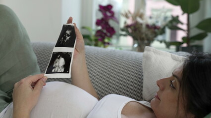 Pregnant woman looks at ultrasound image of her baby during late stage pregnancy laid on couch...