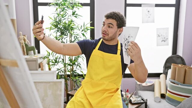 A young man in a yellow apron takes a selfie while holding a sketch in an artist's studio, showcasing creativity and modern technology.