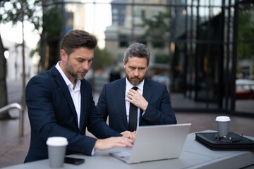 Two businessmen on the street, deeply engrossed, eyes fixed on their laptop screen, perhaps...