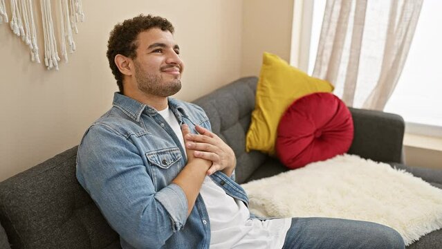 Relaxed young arab man, soaking in the comfort of home, sitting on his trusted sofa, hand on chest, eyes shut, all smiles with a grateful heart and radiant health, basking in a room full of cheer!