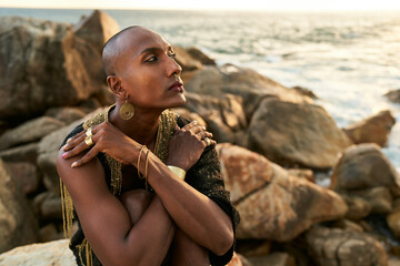 Queer black person in luxury dress, jewelry sits on rocks in ocean. Lgbtq ethnic fashion model...