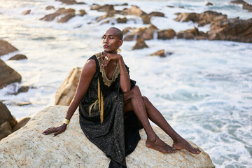 Non-binary ethnic fashion model in dress, brass jewelry sits on rocks by ocean. Trans sexual black person with rings, nose-ring, bracelets, earrings in posh clothes poses in tropical seaside location.