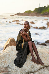 Queer black person in luxury dress, jewelry sits on rocks in ocean. Lgbtq ethnic fashion model...