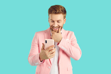 Astonished stylish young man holding smartphone and looking at it. Portrait og handsome man in trendy pastel pink suit using mobile phone isolated over light blue background