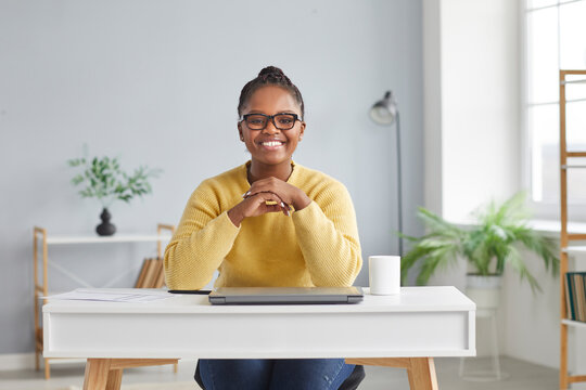 Portrait of happy businesswoman at working desk at home. Beautiful young African American business woman in yellow jumper and eyeglasses sitting at table with laptop, looking at camera and smiling