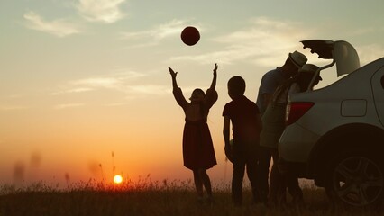 Playful family at sunset by car. cheerful children parents going on travel road trip by automobile packing baggage putting luggage in trunk kids playing ball. Packing preparation for family picnic. - obrazy, fototapety, plakaty