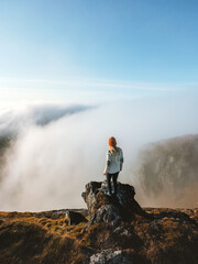 Woman standing on mountain cliff edge above clouds in Norway, traveler hiking solo outdoor active...