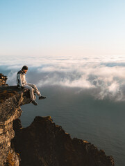 Man traveler sitting on cliff edge of mountain above clouds in Norway, brave tourist hiking solo in mountains outdoor active healthy lifestyle adventure summer vacations