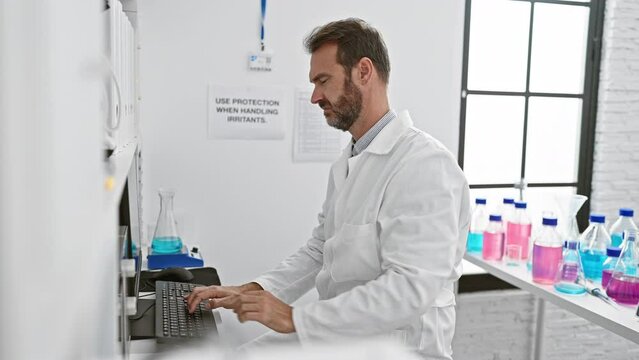 A middle-aged man with a beard works on a computer in a bright laboratory setting surrounded by colorful chemical bottles.