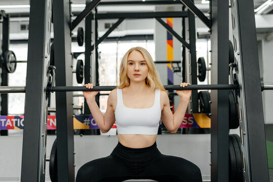 young athletic caucasian woman trains in fitness gym, squats with barbell in smith simulator, blond girl in white top and black leggings, healthy lifestyle concept