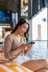 Asian woman with sunglasses sitting and using phone at a poolside bar