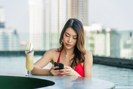 Young woman enjoys sun-kissed serenity, relaxing with drink and using phone in rooftop infinity pool