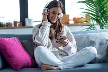 Smiling young woman listening to music with smartphone while sitting on couch at home.