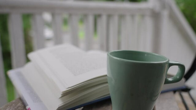 A coffee mug and an open book on a veranda on a breezy, rainy day with cars passing in the background