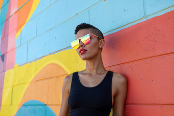 A woman wearing sunglasses stands confidently against a vibrant, multicolored wall, creating a striking visual contrast. - 783369851