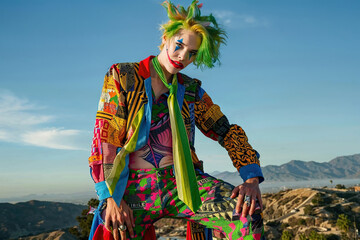 A man in clown attire sitting on a large rock outdoors. - 783369885
