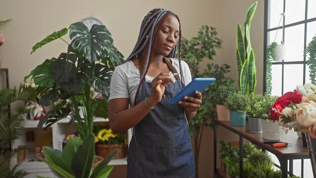 A focused black woman with braids uses a tablet inside a lush flower shop filled with green plants and colorful blooms.