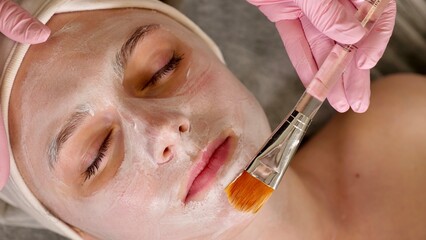 professional cosmetologist applies a white mask on the face of a female client with a brush, close-up