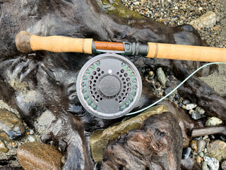 Fly fishing rod and reel on a piece of dark driftwood with riverbank