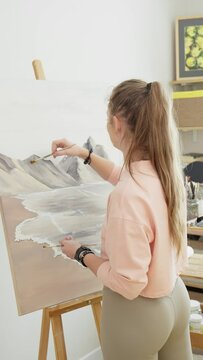 Young stylish female artist painting picture on canvas using acrylic paints. Canvas stands on easel. Female artist works in studio. Creative hobby as leisure and recreation. Vertical footage