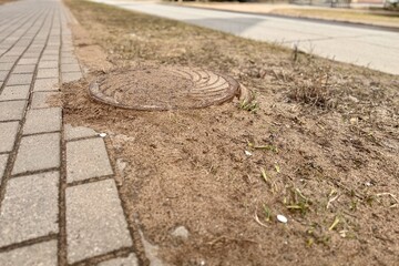 A rusty iron hatch covered grains of sand and the ground with a pattern above an observation well is located next to a dry grass in the daytime. Close-up