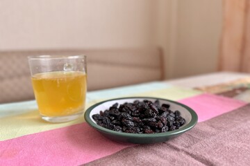 A lot of black raisin in the white green plate and transparent glass of orange juice are located on the table at home