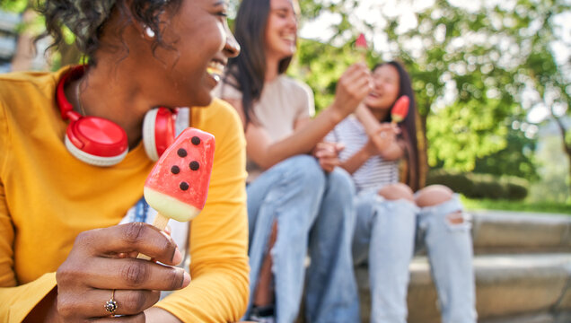 Three young multi-ethnic laughing women sitting eating ice cream in park. Female friends chatting cheerful enjoying summer afternoon together. Focus on black girl in foreground, unfocused background 