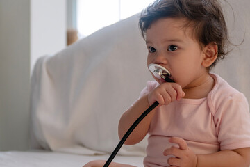 Healthy lovely infant girl playful sucking stethoscope in mouth sitting on bed looking something at...