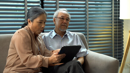 Grandmother pointing finger touchscreen picture on digital tablet for husband Alzheimer’s pain sitting together sofa at home.Elderly couple  memory disability patient health support care. Family care