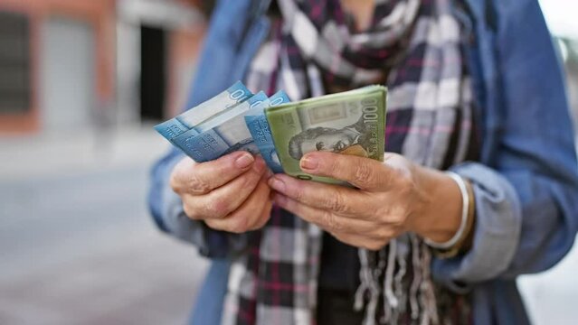 A mature woman examines chilean peso banknotes in her hands against a blurry urban street background.